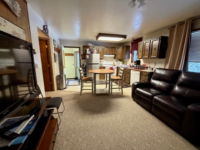 An interior view of a multi-purpose living space featuring a dark leather sofa in the foreground and a small wooden dining table with two chairs in the center. In the background, there is a compact kitchen with dark wood cabinets, a white stove, and a stainless steel refrigerator, alongside a doorway leading to another room.