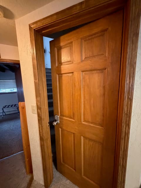 An interior view of a hallway featuring a wooden six-panel door slightly ajar, revealing a carpeted staircase in the background. To the left, a doorway opens into another room with a window, while the surrounding walls are a neutral off-white.