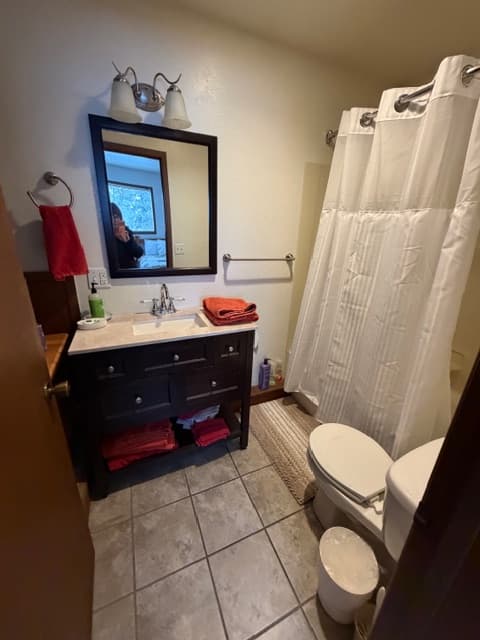 An interior view of a bathroom featuring a dark wood vanity with a white countertop and a rectangular mirror above it. The room includes a white toilet, a bathtub with a white shower curtain, and tan floor tiles. Red towels are neatly placed on the vanity and hung on a wall-mounted ring.