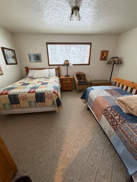 An interior view of a bedroom featuring two beds with different patchwork quilts: one with a colorful block pattern and another with blue and brown patterns. The room includes a wicker armchair, a wooden nightstand with a forest-themed lampshade, and several framed pictures on the walls. A large window provides a view of a snowy landscape.
