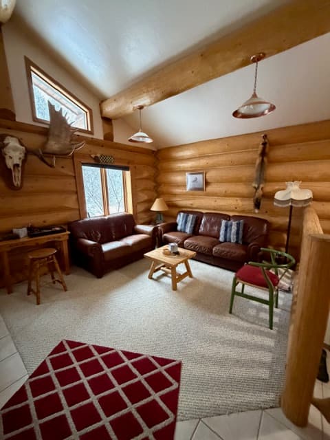 An interior view of a bedroom featuring a bed with a colorful patchwork quilt in a rectangular block pattern of red, brown, tan, and blue. The room includes a light-colored headboard, a tall wooden armoire, and a window providing a view of a snowy outdoor scene. The walls are neutral-toned with wood paneling on the lower half, and a framed landscape picture is mounted above the bed.