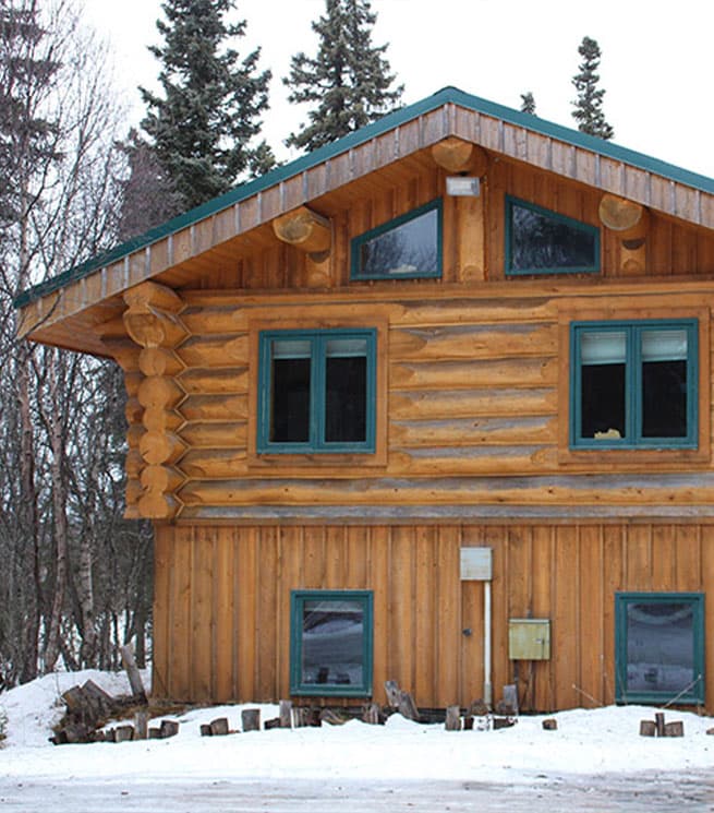 A wooden log house with a sloped roof surrounded by snow and trees.
