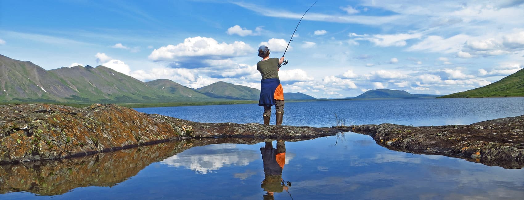 A person stands on a rocky shore casting a fishing line into a calm lake surrounded by mountains and blue skies.