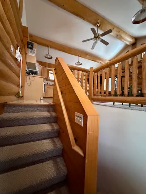 A carpeted staircase with a wooden handrail leads up toward a living area in a rustic log cabin. The upper level features high, vaulted ceilings with large exposed log beams, a ceiling fan, and wooden railings overlooking the stairs.
