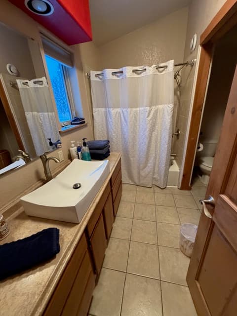 A bathroom featuring a large, modern white vessel sink on a tan countertop with wooden cabinetry below. The room has light-colored floor tiles and a vaulted ceiling. A white shower curtain is partially visible, and a window shows a glimpse of a blue-toned outdoor scene.