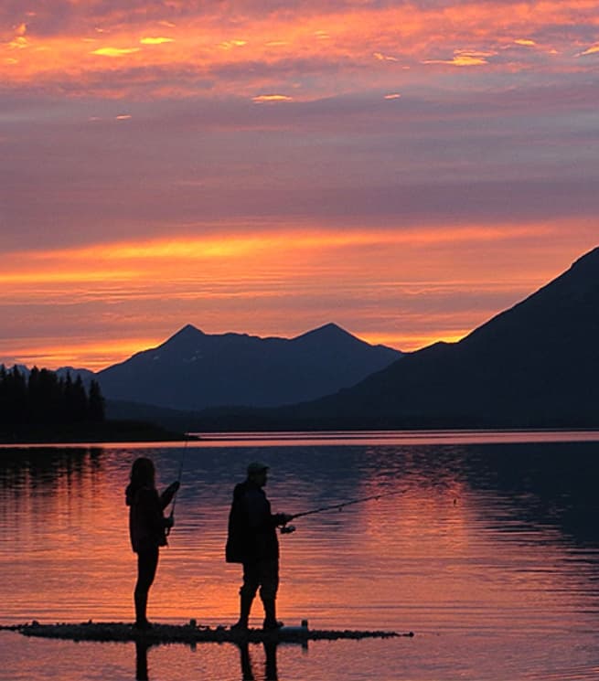 Silhouettes of two fishermen stand by a calm lake at sunset, with mountains in the background.