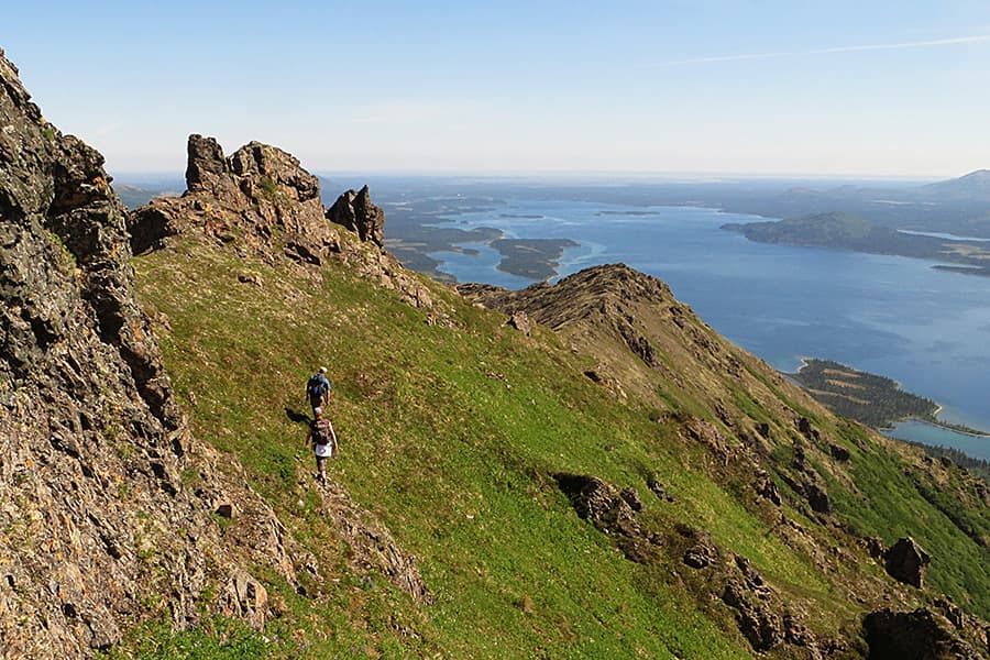 Two hikers traverse a grassy mountain slope with a view of a lake and islands below.