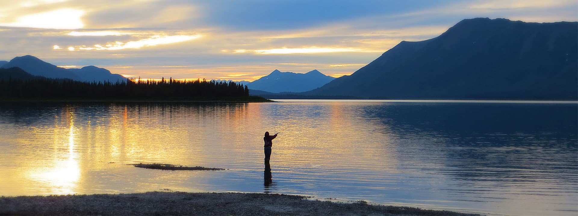 A person stands in shallow water, silhouetted against a vibrant sunset and mountains in the background.