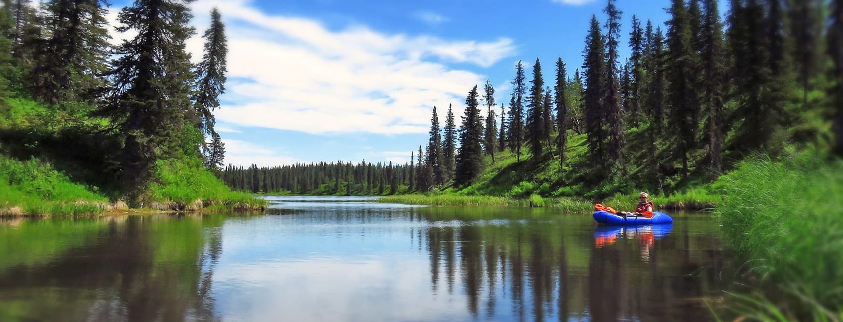 A person in a blue raft paddles on a serene river surrounded by lush greenery and tall trees under a bright blue sky.