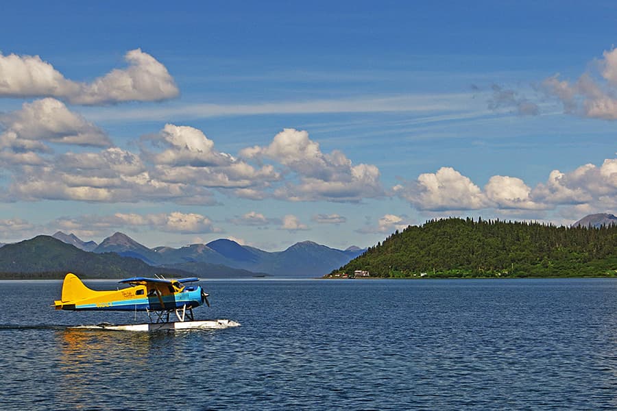 A yellow seaplane glides across a calm lake with lush green hills and mountains in the background under a blue sky with clouds.