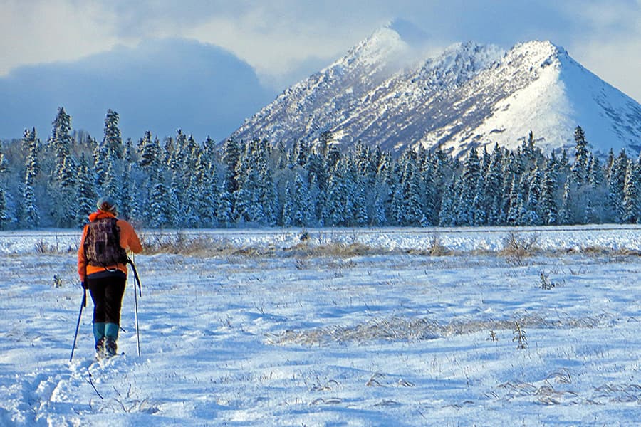 A hiker wearing an orange jacket walks through a snow-covered landscape with mountains in the background.